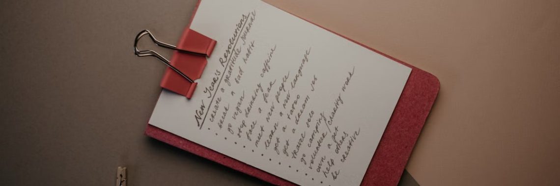 Top view of notebook with handwritten New Year's resolutions, pen, and clips on a desk.